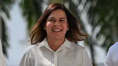 Sara Duterte smiling in a white button-up shirt while standing in front of palm trees