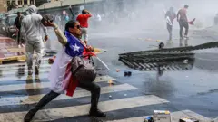 Protesto social de pescadores no Chile. Uma pessoa coberta com a bandeira chilena lança uma grande pedra contra a polícia.