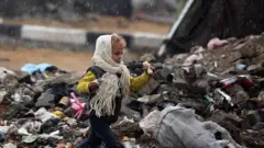 Un niño con bufanda y gorro corre entre un vertedero de desechos