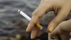 Foto de uma mão com unhas amareladas segurando um cigarro.