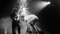 Guitarista inclinando instrumento ao lado de Cazuza agachado, se movimentando no palco