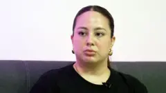 Mulher jovem com cabelos escuros presos para trás, usando brincos de ouro, sentada em um sofá cinza.