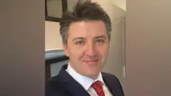 佐尼身穿深色西裝，並且微笑望向鏡頭。