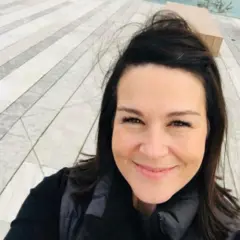 Amy é uma mulher branca, com cabelos castanhos escuros. Ela usa uma roupa preta e sorri para a foto