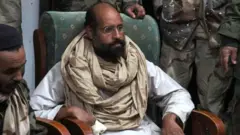 Seyfülislam Kaddafi, Libya'daki ayaklanma sırasında isyancılarca Obari'de yakalanmıştı, 19 Kasım 2011. Fotoğrafta geleneksel kıyafetleriyle, etrafı isyancı savaşçılarla çevrili bir şekilde oturuyor, sağ elinin parmaklarında bandaj var. 