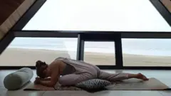 Mulher em posição de ioga em frente a janela de frente para o mar
