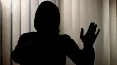 تصویر کې یوه ځپل شوې مېرمن ښکاري چې مخ یې د کوټې پردې پلو دی او تصویر له شا لورې اخېتسل شوی دی. 