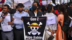 Un hombre con una mascarilla negra sostiene un cartel negro con una calavera y huesos cruzados y las frases "Gen Z" y "Nepo Babies" impresas, el 8 de septiembre de 2025. Hombres y mujeres se encuentran a su lado, con una gran multitud reunida al fondo. Un cartel dice "Protejamos nuestro Nepal".