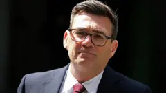Andy Burnham, nuevo primer ministro de Reino Unido. Tiene el pelo castaño corto, con anteojos, una chaqueta oscura y corbata roja