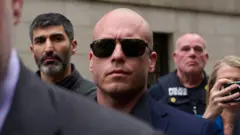 Um homem branco, careca, de óculos escuros e terno azul-marinho faz expressão séria. Ele está cercado por várias pessoas, entre elas um policial
