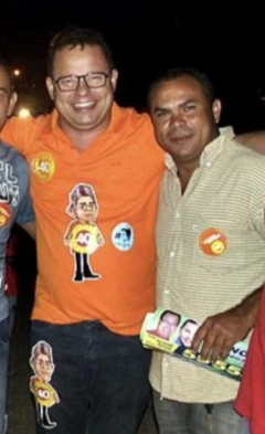 Dois homens se abraçam e olham para a câmera, com materiais de campanha em suas camisetas e um panfleto na mão de um deles