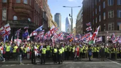 Uma multidão de centenas de pessoas se reúne no cruzamento de duas ruas de Londres, com altos arranha-céus da City visíveis ao fundo. Há dezenas de bandeiras do Reino Unido e da cruz de São Jorge. À frente da multidão está uma fileira de policiais com jaquetas de alta visibilidade.