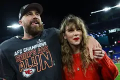 Taylor Swift e Travis Kelce em um jogo de futebol americano. O braço dele está sobre o ombro dela e ela segura o dedo médio dele com a mão esquerda.
