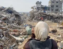 Uma mulher de véu claro e casaco cinza e marrom aparece de costas. À sua frente, restam apenas as estruturas de dois prédios destruídos e grandes montes de entulho e terra.