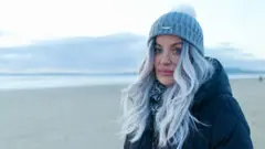 Joanne, who has trendy dyed gray hair and blue eyes, wears a gray woolen hat and black coat, looks seriously at the camera on a vacant Welsh beach at dusk at dusk