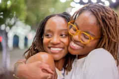 Duas irmãs gêmeas afro-americanas sorrindo, na casa dos vinte anos, abraçando-se carinhosamente