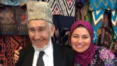 زینب دست راستش را با بر شانه پدرش حلقه کرده است. او پیراهنی گلدار بر تن دارد و حاجی محمدعوض بادغیسی کلاه پوست سنتی بر سر دارد. در پسزمینه نیز پارچههای رنگارنگ و نقشدار افغانستان و ازبیکستان دیده میشود.