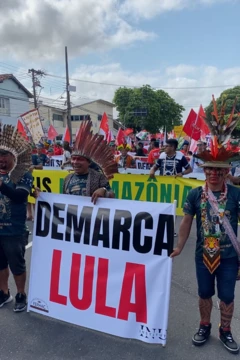 Manifestantes indígenas vestindo adereços tradicionais e segurando um cartaz onde se lê "Demarca Lula"
