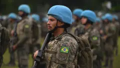 Um militar brasileiro usando uniforme camuflado, capacete azul e mochila aparece em primeiro plano segurando uma arma, enquanto outros militares com capacetes azuis permanecem perfilados ao fundo, desfocados.
