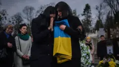 A mãe de um soldado chora enquanto abraça a bandeira ucraniana durante o funeral de três soldados mortos em decorrência dos ataques russos na cidade de Mykolaiv, no Cemitério Lychakiv, em Lviv, na Ucrânia, em 29 de março de 2022.