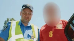 Temerbek aparece en la imagen con una gorra policial negra, una camisa de policía azul y un chaleco amarillo de alta visibilidad, mirando a la cámara con una leve sonrisa. A su lado se encuentra una persona con una camiseta de fútbol roja, cuyo rostro ha sido difuminado.