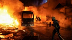 Üç itfaiye görevlisi alev alev yanan bir otobüsü su sıkarak söndürmeye çalışıyor. Gece karanlığında ortam alevlerin ışığıyla aydınlanmış durumda.