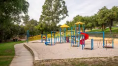 誰もいない公園。砂場の上に赤色の滑り台などの遊具がある。周囲には木々が点在し、緑地が広がっている
