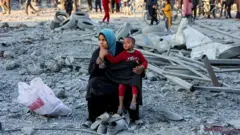 一名婦女帶著兒童坐在灰色廢墟中，若有所思。