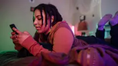 Una estudiante en la cama boca abajo con un teléfono celular en la mano. 