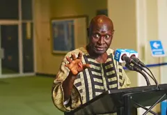 Olara Otunnu, umukandida wa Uganda ku mwanya w’umunyamabanga mukuru wa ONU, aganira n’abanyamakuru ku wa gatatu ku cyicaro gikuru cya ONU i New York, muri Amerika, ku itariki ya 19 Kanama (8) mu 2026, nyuma y’ikiganiro cyo ku ruhande yagiranye n’ibihugu binyamuryango bya ONU n’imiryango inyuranye. Yambaye ishati nini y’igitenge y’amabara y’umuhondo n’umukara, akoze ikimenyetso n’ikiganza kimwe. Arimo kuvugira mu ndangururamajwi ebyiri zegeranye zanditseho amagambo ‘UN’.