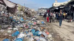Deslocados da Faixa de Gaza caminham por um mercado ao lado de um lixão repleto de sacolas plásticas. Um homem passa com uma criança nos ombros, ambos olhando fixamente para o lixão. Vários homens, mulheres e crianças o seguem. Vans brancas e prédios em ruínas são visíveis ao fundo.