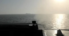 Un barco de carga en el horizonte visto desde la cubierta de otro barco del que sale humo. 