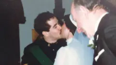 Há quatro pessoas nesta fotografia. No centro, um casal de noivos se beija no dia do casamento. O noivo veste um uniforme militar preto. A noiva, de costas para a câmera, usa um vestido branco de gola alta e um véu branco preso a um adorno de cabeça. Ambos têm cabelos escuros. À esquerda do noivo está o cônego, que celebrou o casamento. À direita da noiva, um convidado do casamento veste um terno preto.