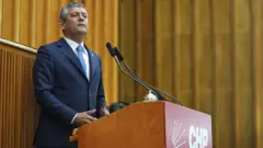 Özgür Özel CHP logolu bir kürsüde konuşma yapıyor. Üzerinde lacıvert bir takım var.