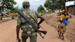 Ifoto yo mu bubiko igaragaza umurwanyi wa PARECO mu gice cyo mu mugongo, ahetse imbunda afite n'amasasu mu ntoki, iruhande rwe hari umugore uhetse umwana. 