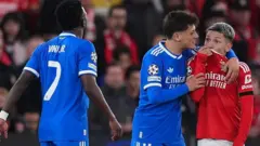 Gianluca Prestianni, com uniforme vermelho, cobre a boca com a camisa enquanto se dirige a Vinícius Jr., com uniforme azul, que está à sua frente. Prestianni é abraçado pelo companheiro de equipe de Vinícius Jr., Arda Güler.
