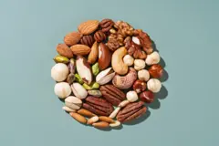 Mistura de nozes que inclui amêndoas, castanhas-do-pará, castanhas de caju, avelãs, macadâmia, nozes-pecã e pistaches, dispostas em círculo sobre um fundo verde, vistas de cima