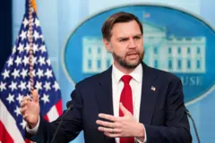 Fotografia colorida mostra o vice-presidente dos Estados Unidos, JD Vance, um homem branco de barba e olhos claros, usando um terno escuro e gravata vermelha. Ele gesticula em frente a microfones. Atrás dele, há uma bandeira americana e o símbolo da Casa Branca.