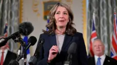 La primera ministra de Alberta, Danielle Smith, habla frente a varios micrófonos. Viste una camisa gris y una chaqueta azul oscura
