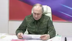 Poutine est assis à une table et lit une feuille de papier écrite à la main. À l'arrière-plan se trouve une peinture aux couleurs rouge et violette. Il porte une tenue de combat