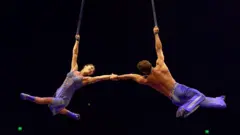 Dois artistas do Cirque du Soleil, uma mulher e um homem com figurinos roxos combinando, dão as mãos enquanto balançam em um trapézio durante um espetáculo realizado em 08/2025, em Perth (Austrália)