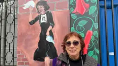 Une femme, Alejandra Holzapfel, se tient devant l'un des murs extérieurs peints du site commémoratif qu'elle dirige, anciennement connu sous le nom de « Venda Sexy », un ancien centre secret de détention et de torture pendant la dictature d'Augusto Pinochet, aujourd'hui connu sous son adresse actuelle Iran 3037. La femme regarde droit dans les yeux la caméra et sourit. Elle a des cheveux roux courts et plumeux, des lunettes de soleil noires à bords gris et porte des boucles d'oreilles ornées, un pull à col roulé noir et un cardigan en laine tricoté violet, rose et gris. Derrière elle, il y a un tableau représentant une femme en noir tenant un mouchoir blanc, alors qu'elle regarde le sol avec tristesse. Elle a la photo d'une personne disparue collée sur sa poitrine.