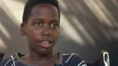 ザヘルはカメラを見て話しています。少年は車椅子に座っており、白い縞模様の着古された黒いTシャツを着ています。