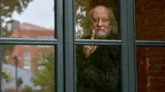 الكاتب الهنغاري لازلو كراسنهوركاي يقف خلف نافذة زجاجية، يرفع إصبعه بهدوء وينظر بتأمل إلى الخارج، فيما تنعكس خلفه ملامح مبانٍ من الطوب وأشجار خريفية.