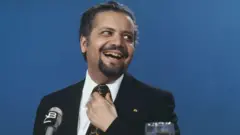 Ahmed Zaki Yamani frente a um microfone, rindo