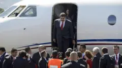 A foto mostra Maduro, um homembwin hrvatskapele clara, cabelo e bigode pretos, vestindo um terno escuro sobre camisa branca e gravata vermelha, descendo da escadabwin hrvatskaum avião. 