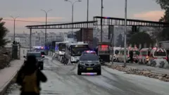 Çakarlı bir araç önde arkadan otobüsler geliyor. Geniş yolun sağında ambülanslar park etmiş. 