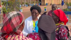 Nathalie Mukamazimpaka ari imbere y'abagore batatu bambaye ibitambaro mu mutwe, afite imisatsi myinshi, yambaye n'umwenda w'umweru