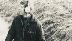 Una imagen en blanco y negro de Dave Derby, que data de la década de 1970 aproximadamente. Mira hacia la izquierda de la imagen y lleva puestos unos auriculares con cable y una chaqueta medio abrochada sobre un jersey de cuello alto. Detrás de él se ve el borde cubierto de hierba de una zanja.