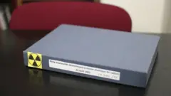 Caixa pequena, na cor cinza, está em cima da mesa. Na lateral há um símbolo amarelo, alertando para a radioatividade